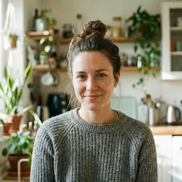 SGO-Immo-Simon-Gogel- Eine Frau mit braunen Haaren in einem Dutt und einem grauen Pullover sitzt in einer stilvollen Reutlinger Küche mit Regalen, Pflanzen und Küchenutensilien. Sie blickt in die Kamera und lächelt leicht, um einen warmen Moment in ihrer von Immobilien gestalteten Wohnung festzuhalten.