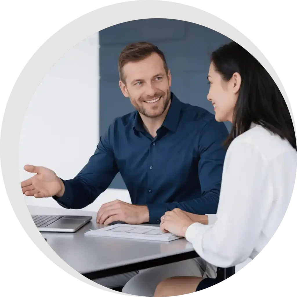 SGO-Immo-Simon-Gogel- Ein Mann in einem blauen Hemd und eine Frau in einer weißen Bluse sitzen an einem Schreibtisch in Reutlingen, unterhalten sich und lächeln. Der Mann gestikuliert, während sie über die Instandhaltung von Immobilien diskutieren, während Papiere und ein Laptop auf dem Tisch liegen.